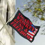 Flag with 'Cigars, Guns & Whiskey Freedom' text and American flag design on a pole against a building and tree background.