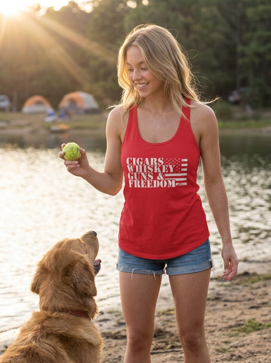 A woman standing outdoors by a lake at sunset wearing a red racerback tank top with the text 'CIGARS WHISKEY GUNS & FREEDOM' and a distressed American flag graphic. She is holding a green tennis ball and looking at a golden retriever dog.