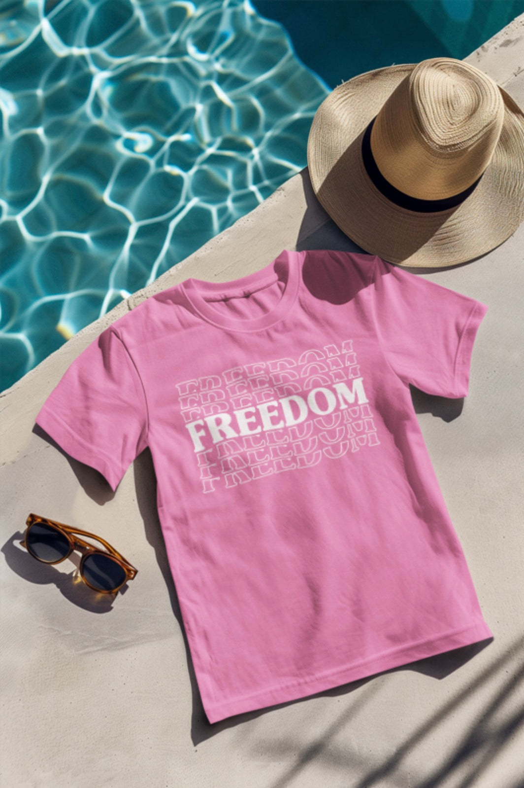 Pink t-shirt with 'FREEDOM' text, sunglasses, and straw hat by a pool.