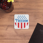 Coaster with 'Freedom' design on a wooden surface with a drink and notebook.