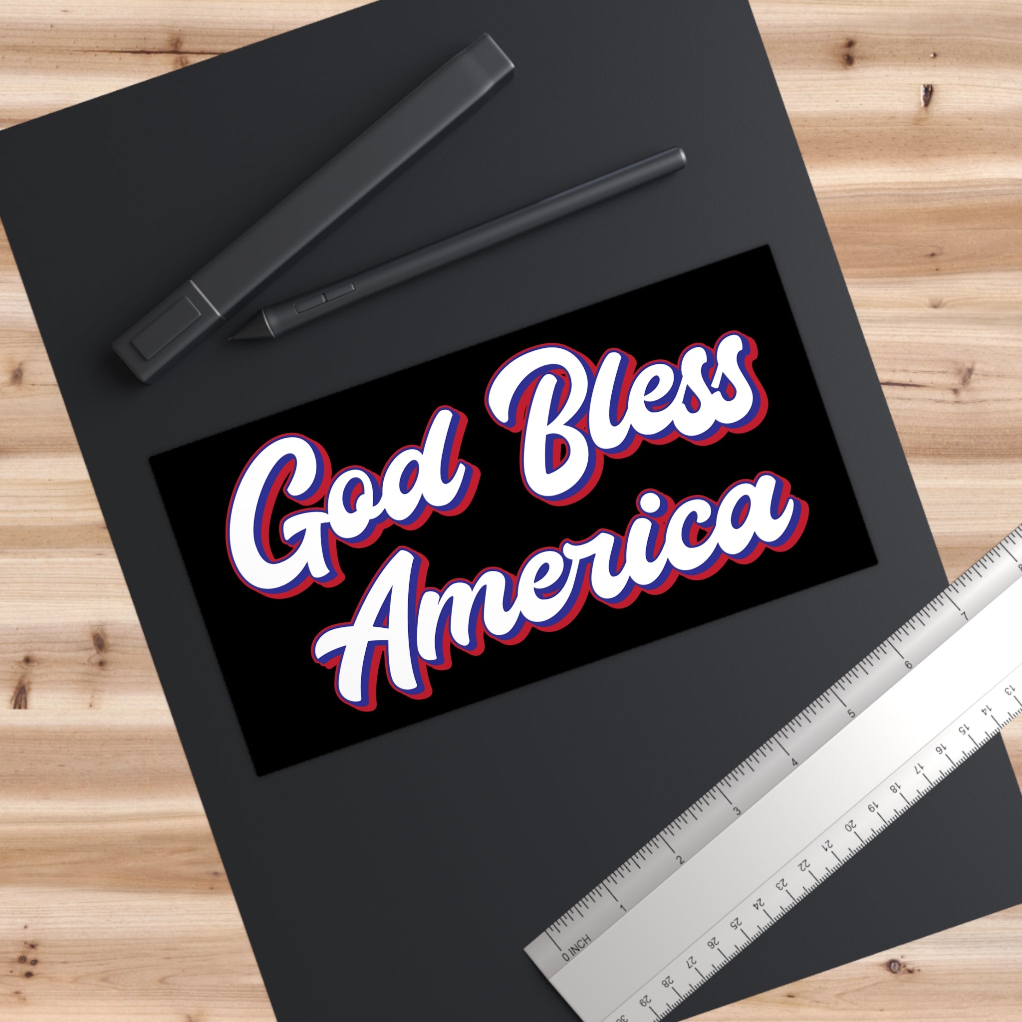 Black frame with a Vinyl Bumper Sticker with 'God Bless America' text on a wooden surface with pens and a ruler.