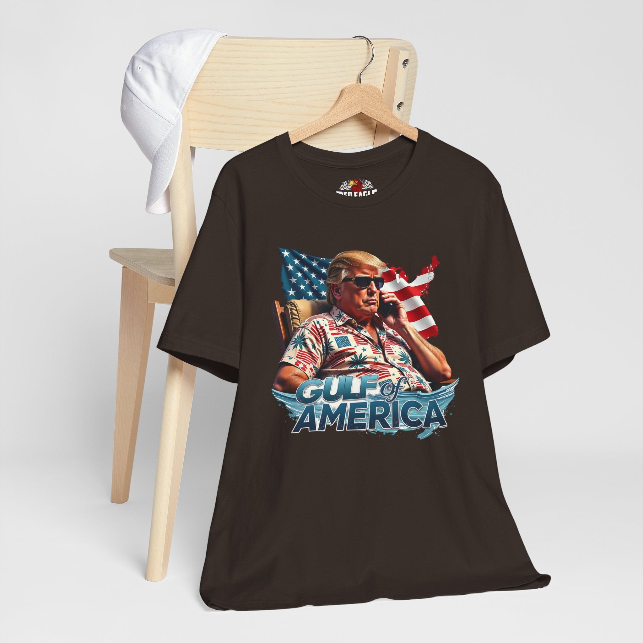 Brown t-shirt with graphic design of a person holding a phone in front of an American flag, sitting in a chair, with 'Gulf of America' text on a white background.