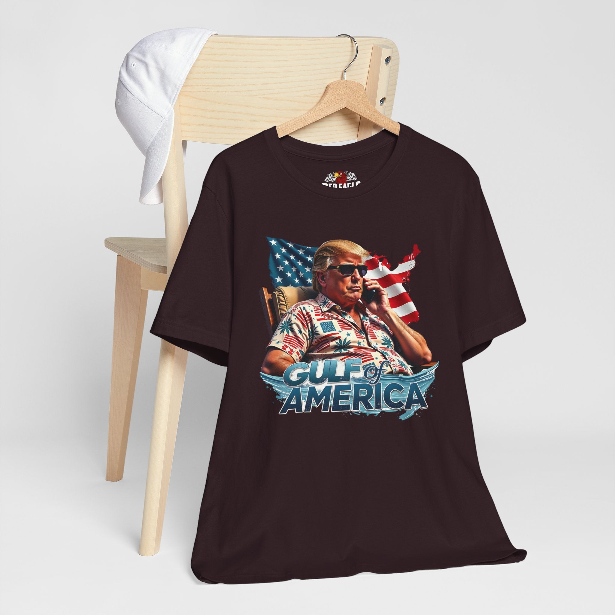 Oxblood Black t-shirt with graphic design of a person holding a phone in front of an American flag, sitting in a chair, with 'Gulf of America' text on a white background.