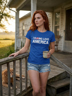 A woman with red hair stands on a wooden porch wearing a royal blue graphic t-shirt that reads 'I FEEL LOVE AMERICA' with a small American flag graphic, paired with denim shorts and holding a coffee mug. A golden retriever lies on the porch steps in the background. (AI generated)
