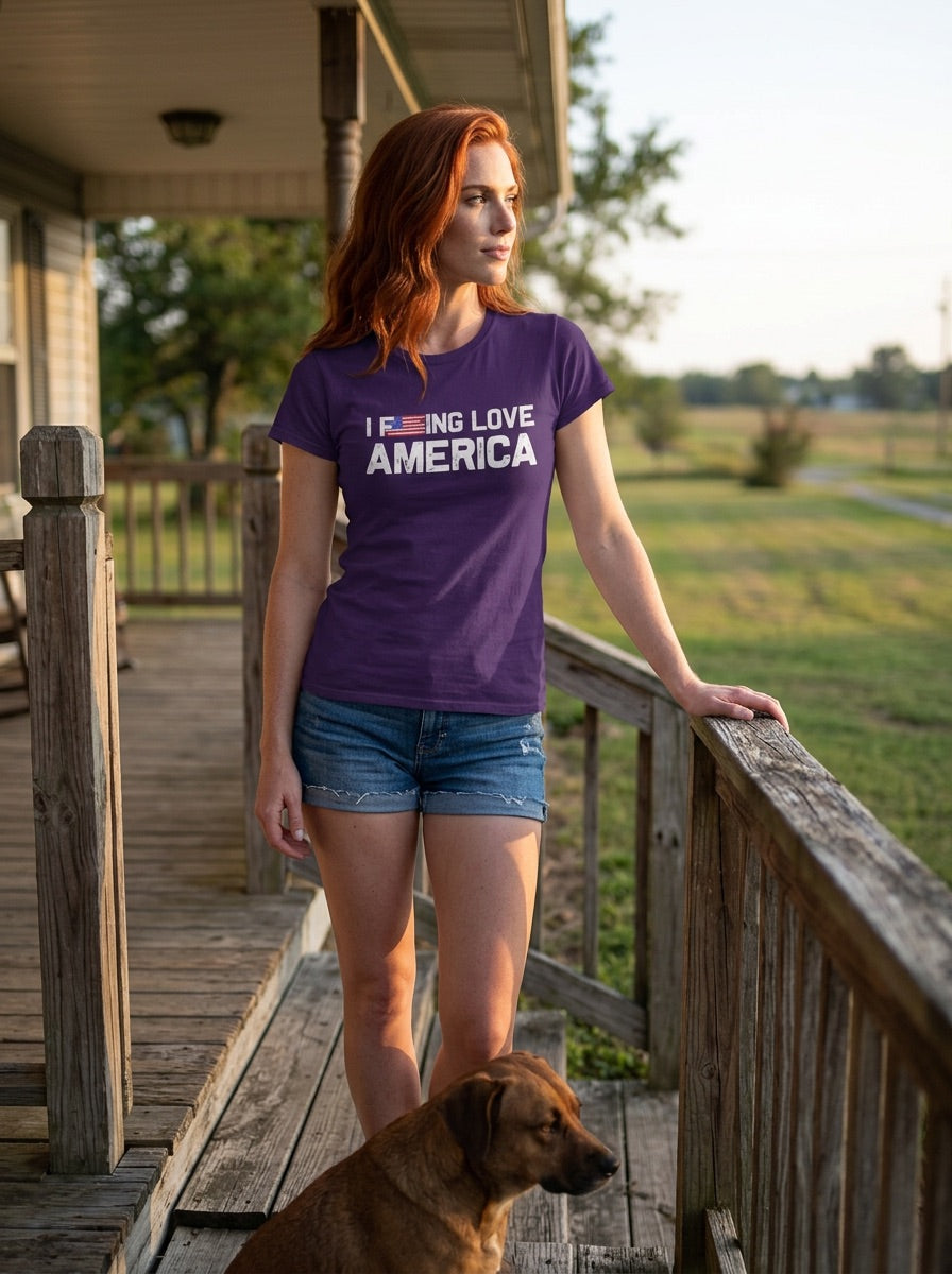 A woman with red hair stands on a wooden porch wearing a purple t-shirt with the text 'I DON'T LIKE AMERICA' in white, paired with denim shorts, with a brown dog at her side and a grassy field in the background.