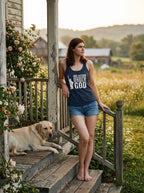 A woman stands barefoot on rustic wooden porch steps wearing a navy blue tank top with the white printed text 'ONE NATURAL UNDER GOD' and denim shorts, with a yellow Labrador retriever lying on the porch beside her in a rural outdoor setting.