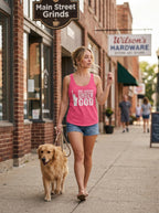 A woman walking a golden retriever on a sidewalk in front of brick storefronts, wearing a bright pink tank top with the white text 'ONE NATION UNDER GOD' and denim shorts.