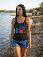 A woman standing on a lakeshore wearing a black racerback tank top with a blue and red graphic that reads 'ONE NATION UNDER GOD' alongside a Statue of Liberty silhouette, paired with denim shorts.