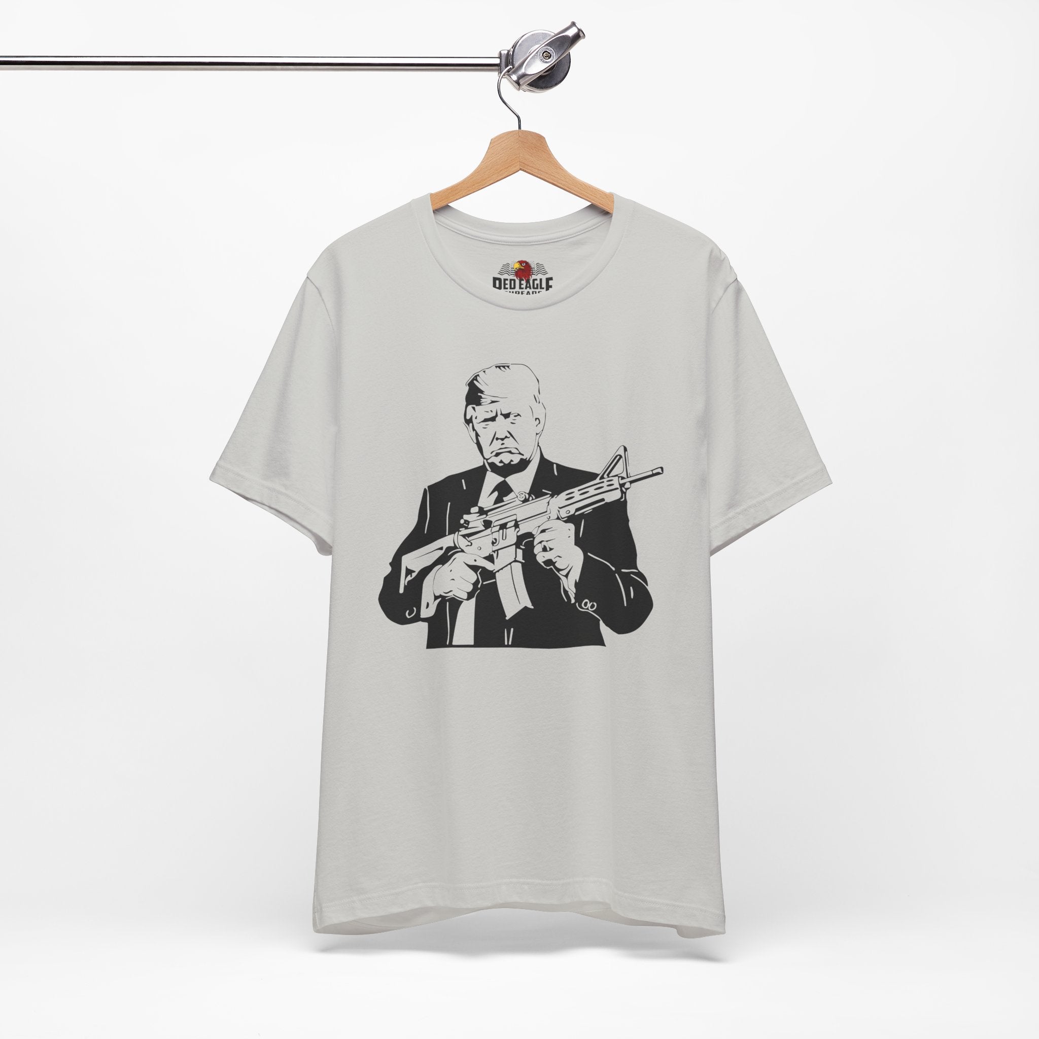 Silver t-shirt with graphic design of a person holding a rifle, hanging on a wooden hanger against a white background.