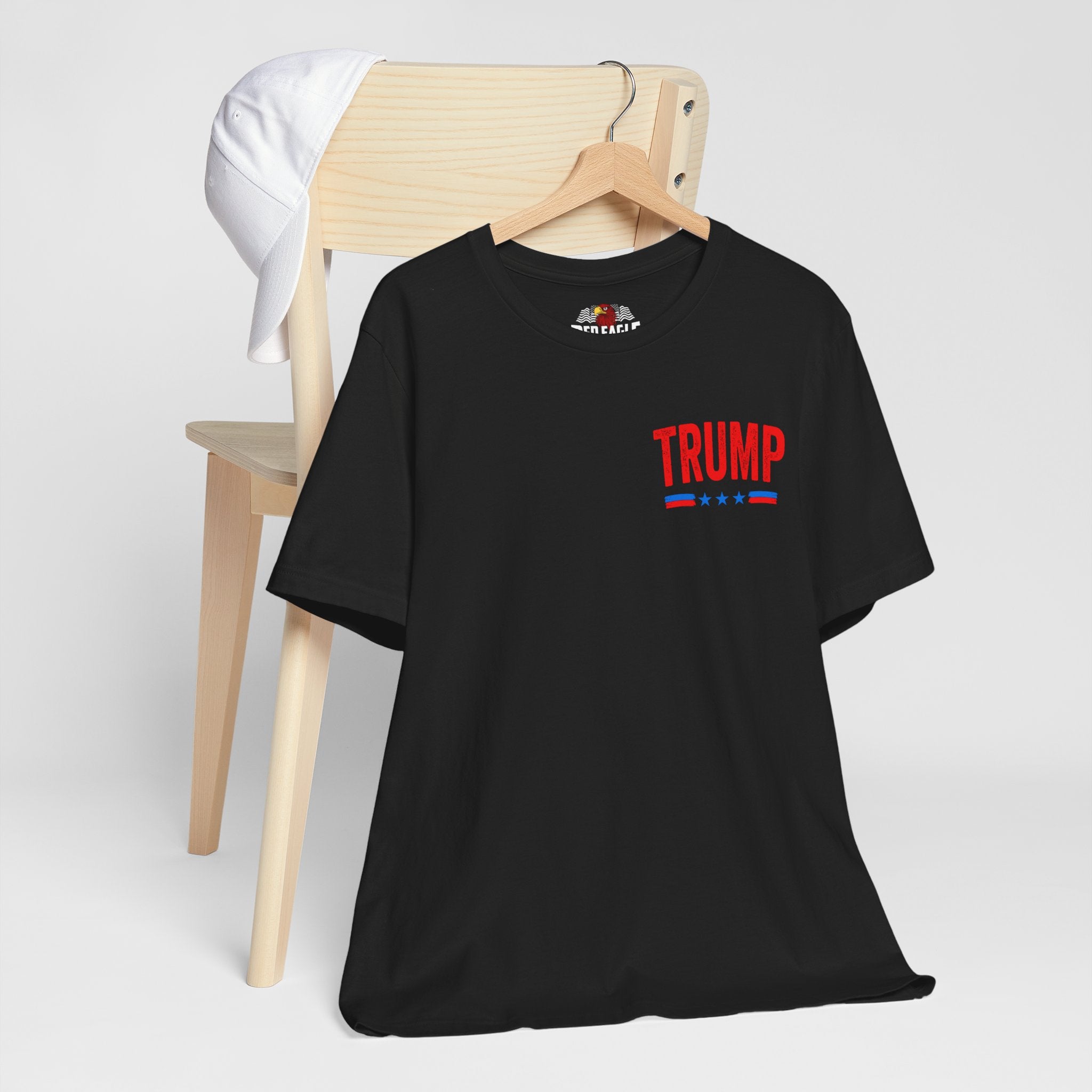 Black t-shirt with 'TRUMP' printed on the front, hanging on a wooden chair against a white background.