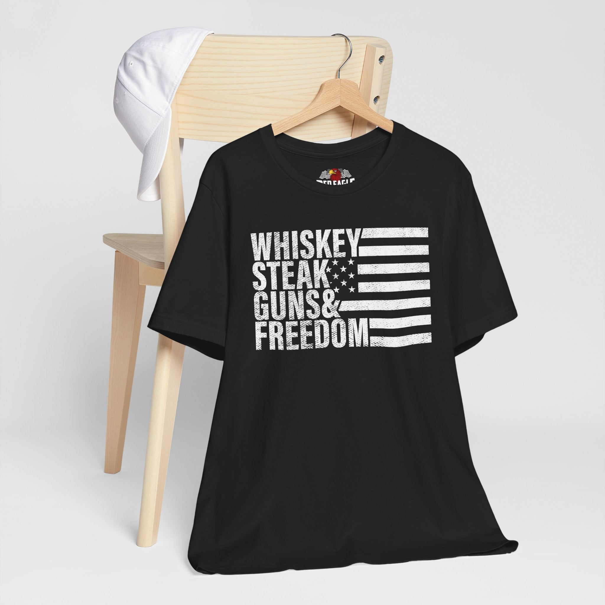 Black t-shirt with 'Whiskey Steak Guns & Freedom' text and flag design on a wooden chair.