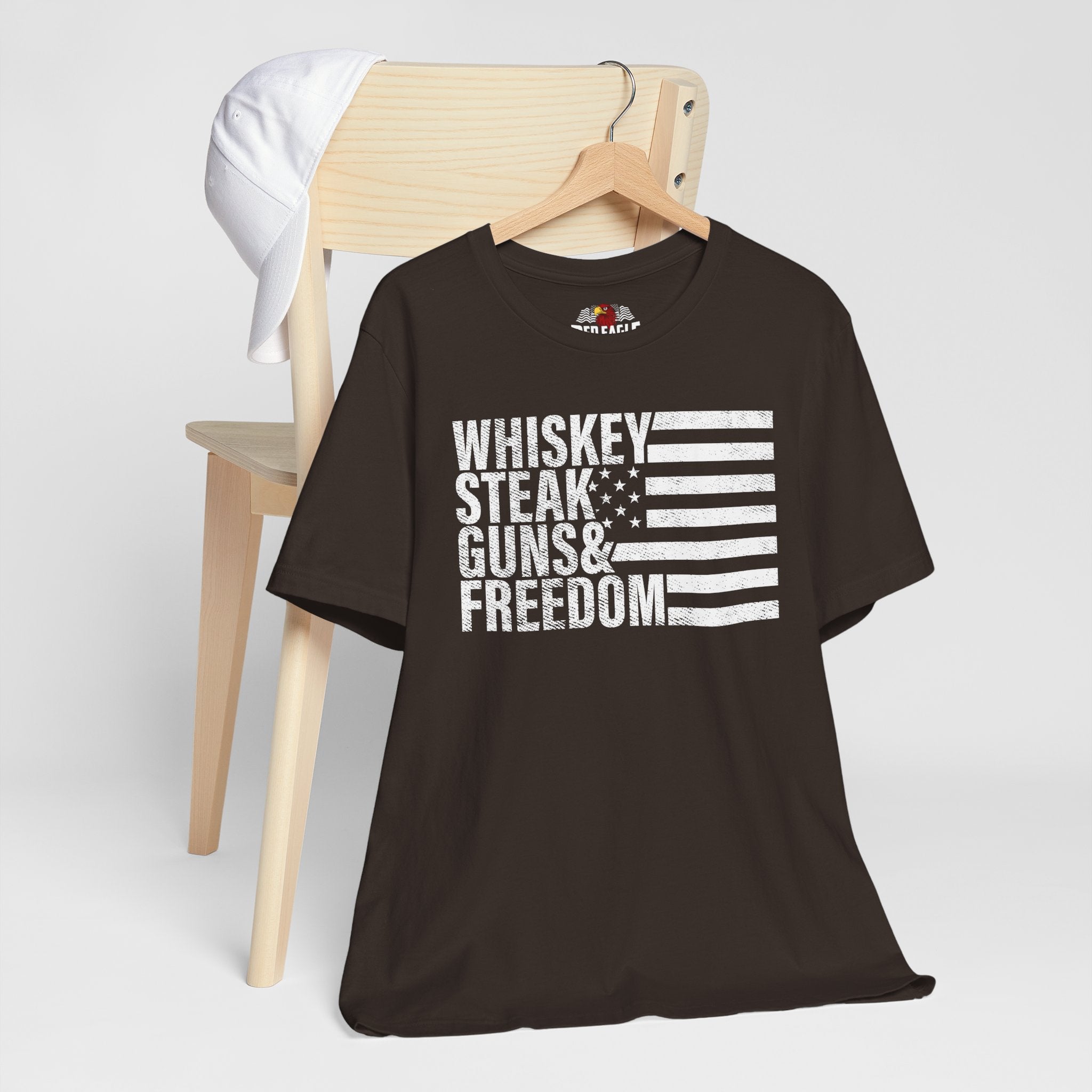 Brown t-shirt with 'Whiskey Steak Guns & Freedom' text and flag design on a wooden chair.