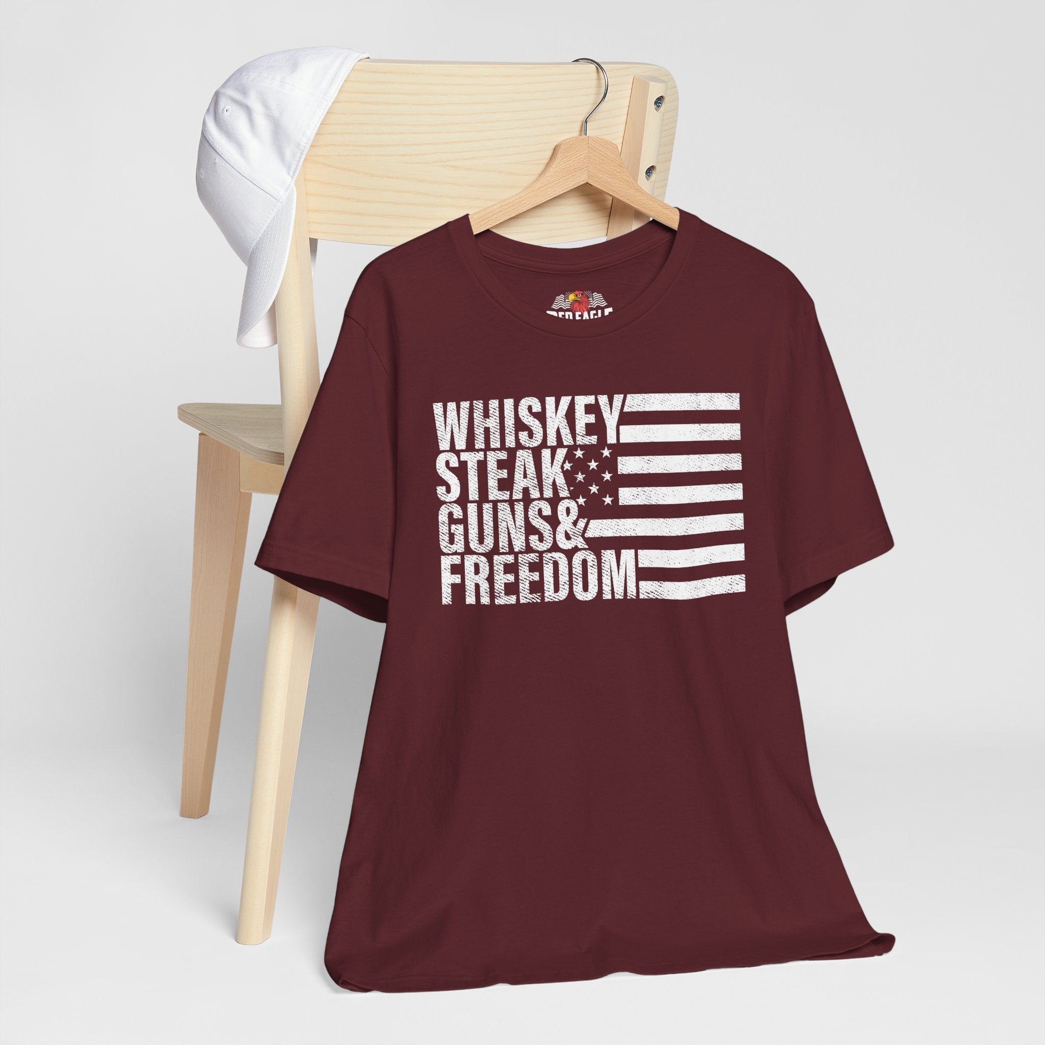 Maroon t-shirt with 'Whiskey Steak Guns & Freedom' text and flag design on a wooden chair.
