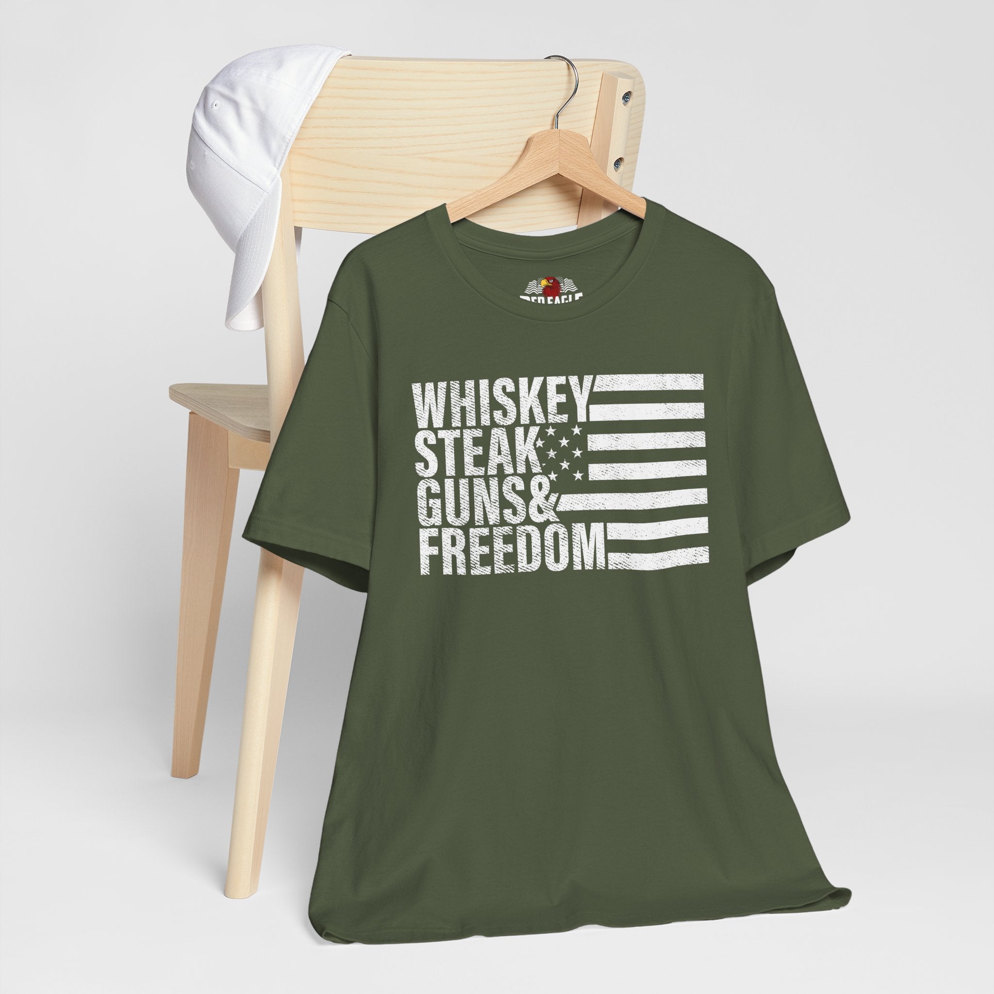 Military Green t-shirt with 'Whiskey Steak Guns & Freedom' text and flag design on a wooden chair.