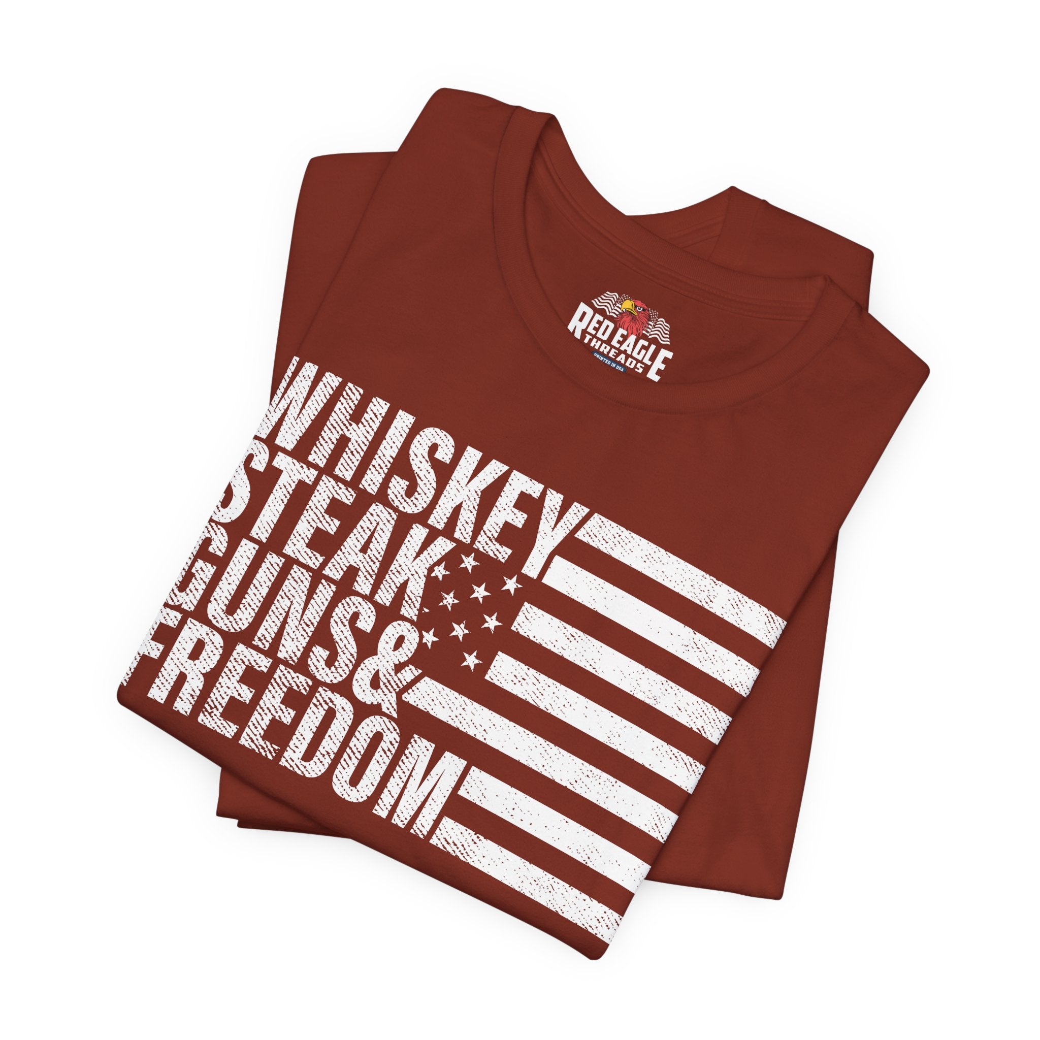 Folded rust t-shirt with 'Whiskey Steak Guns & Freedom' text and American flag design on a white background.
